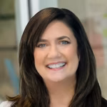 Smiling woman with straight dark brown hair, wearing a light-colored top, standing indoors with a blurred background.
